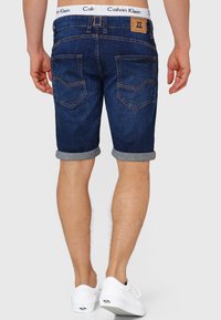 Tamno plavi traper šortsevi s poderanim rubovima, vidljivim šavovima i dva stražnja džepa. Calvin Klein logo na pojasu. Upareni s bijelim cipelama.