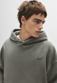 Grauer, übergroßer Hoodie aus weichem Stoff, mit großer Kapuze und kleinem gestickten Logo auf der linken Brust. Glatte Textur.