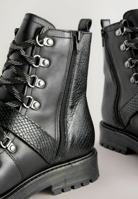 Fekete fűzős bokacsizma strukturált panelekkel, sima bőrből készült felsőrésszel, oldalsó cipzárral és masszív gumitalppal. Fém fűzőlyukakkal és díszítésekkel rendelkezik.
