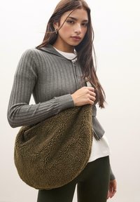 Oversized olijfgroene nep-bont tas met een ronde vorm, zachte textuur en ritssluitingaccent. Model draagt een geribbeld grijs hoodie en zwarte leggings.