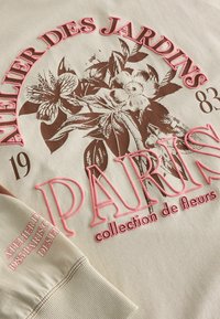 Beige színű pulóver, virágos grafikai elemekkel barna és rózsaszín árnyalatokban. A szöveg tartalmazza az "ATELIER DES JARDINS" és a "PARIS collection de fleurs" feliratokat. A passzé mandzsetták láthatóak.