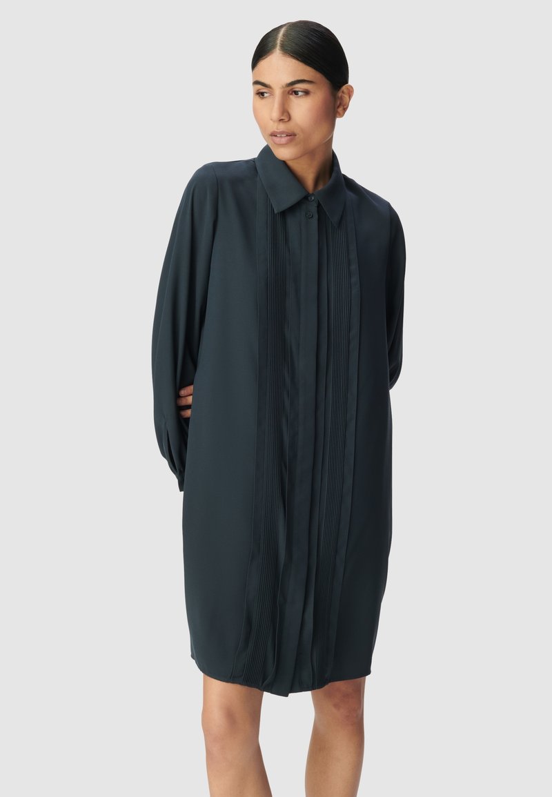 Abito lungo a maniche lunghe di colore verde scuro con colletto a bottone, caratterizzato da pieghe verticali sul davanti e una texture liscia. Forma su misura con vestibilità comoda.