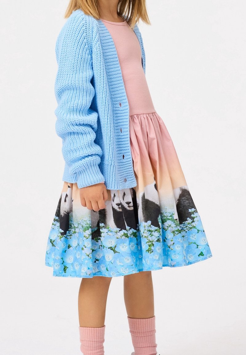 Enfant portant une robe rose avec imprimé panda et fleur bleue, un cardigan bleu clair en tricot et des chaussettes roses sur fond blanc.