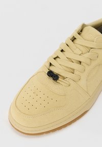 Sneaker gialla in suede con punta perforata, lacci neri piatti e dettagli circolari. La suola in gomma presenta un colore marrone chiaro. Finitura durevole e testurizzata.