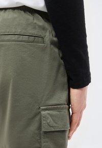 Άτομο που φοράει ελιάς πράσινο παντελόνι cargo και μαύρο μακρυμάνικο τοπ, με το χέρι να ακουμπάει δίπλα στην τσέπη του παντελονιού με λεπτομέρεια καπακιού.