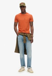 Orange kortærmet t-shirt med lomme, lyseblå jeans, grøn sweatshirt knyttet om livet, beige sneakers og en grøn kasket med logo.