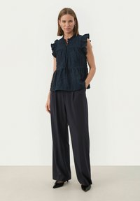 Marineblaue Bluse mit Puffärmeln aus strukturiertem Stoff, gerüschten Akzenten und einem gestuften Saum, kombiniert mit weiten dunklen Hosen und spitzen schwarzen Schuhen.