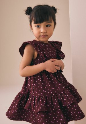 Junges Mädchen mit dunklem Haar, das zu zwei Dutts frisiert ist, trägt ein ärmelloses, burgunderrotes Kleid mit rosa Punkten und steht vor einem schlichten hellen Hintergrund.