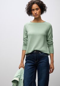 Maglione verde chiaro a maniche lunghe con scollo rotondo e finitura testurizzata. Abbinato a jeans blu scuro, accompagnato da una giacca leggera.