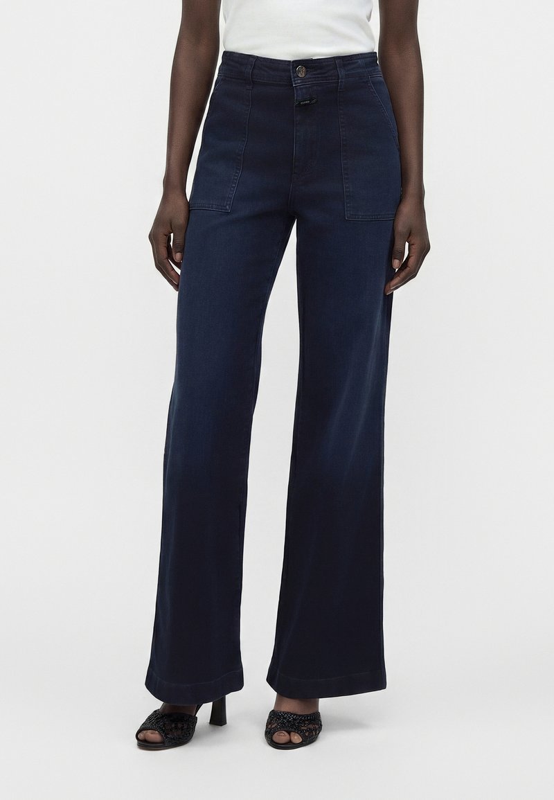 Femme portant un pantalon large bleu foncé avec des poches plaquées à l'avant et des sandales à talons noires à bouts ouverts, debout devant un fond uni.