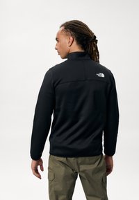 Chaqueta de forro polar negra con cuello alto y logo en la parte posterior, con una textura suave y un diseño ajustado. Combina con pantalones cargo verdes olivo.