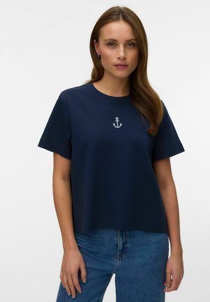 Kvinde med langt brunt hår iført marineblå t-shirt med lille hvid ankeremblem og blå jeans, stående mod en ensfarvet baggrund.