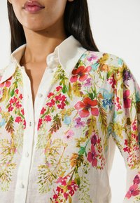 Floral blouse with a white base, featuring colorful patterns of flowers and leaves, a pointed collar, and button closure. Lightweight texture.