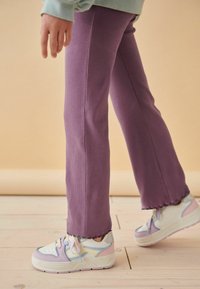 Pantalons lilas côtelés à bas évasés, associés à des baskets blanches avec des accents lilas et bleus, posés sur un sol en bois.