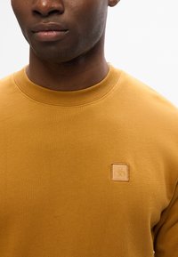 Felpa gialla senape con girocollo, colletto a costine e un piccolo logo quadrato testurizzato sul petto. Tessuto morbido.