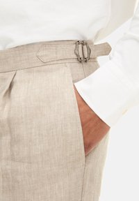 Beige linnebyxor har en skräddarsydd form med ett bälte och metallspänne, kompletterat av en vit knapp-up skjortärm som syns.