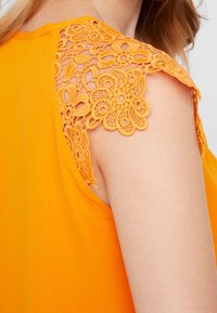 Top arancione senza maniche con un sovrapposto in pizzo sulle spalle decorato con motivi floreali. Tessuto morbido e scollo rotondo.