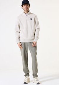 Sudadera gris claro con cremallera y un pequeño logo en el pecho, combinada con pantalones tapered gris claro y zapatillas beige. El fondo es blanco liso.