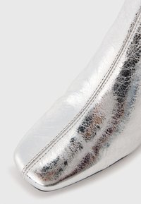 Nahaufnahme der silbernen metallischen Lederbootspitze mit sichtbaren doppelten Nähten und strukturierten Oberflächen auf einem weißen Hintergrund.