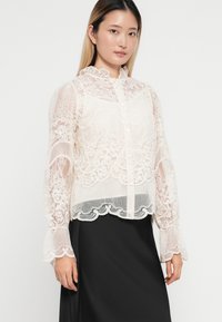 Blusa de encaje en crema con detallados patrones florales, cierre de botones y cuello con volantes. Mangas largas con puños acampanados y dobladillo cropped.