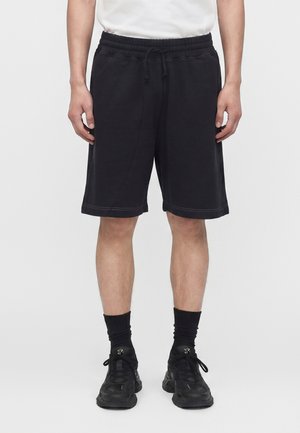 Muž oblečený v čiernych šortkách po kolená, čiernych ponožkách, čiernych teniskách a bielej košeli, stojaci pred jednofarebným pozadím.