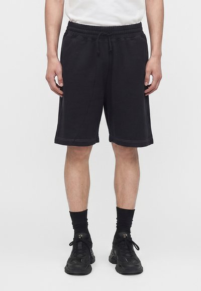 Homme portant un short noir au genou, des chaussettes noires, des baskets noires et une chemise blanche, debout devant un fond uni.