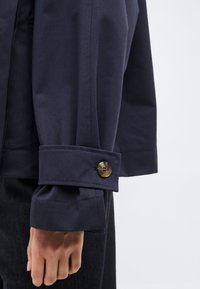 Μανίκι σκούρου ναυτικού σακακιού με μανσέτα με κουμπιά που ακουμπάει σε μαύρο υφασμάτινο παντελόνι, το χέρι χαλαρό δίπλα στο σώμα.