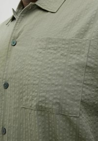 Svijetlozelena košulja na dugme s teksturiranim materijalom, koja ima džep na prsima i tamnozelene dugmadi. Vidljive su vertikalne linije i uzorci.