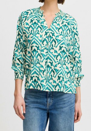 Woman wearing teal and white patterned long-sleeve blouse with gathered cuffs, paired with blue denim jeans.