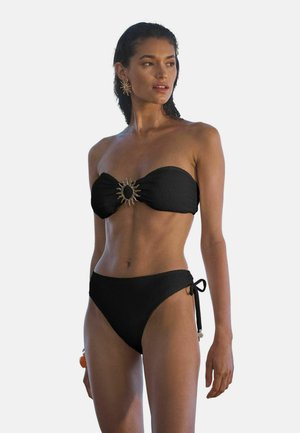 Femeie care poartă un bikini bandeau negru texturat, cu decorațiuni aurii în formă de soare pe partea de sus și slipuri cu șnururi laterale, stând și uitându-se spre dreapta.