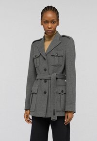 Šedý vlněný kabát s pásem v pase, dvouřadým designem, čtyřmi předními kapsami a ramenními epauletami. Hladká textura.