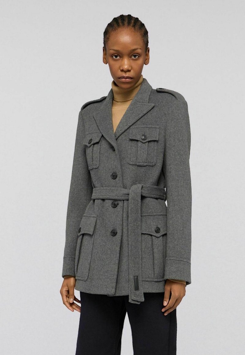 Šedý vlněný kabát s pásem v pase, dvouřadým designem, čtyřmi předními kapsami a ramenními epauletami. Hladká textura.