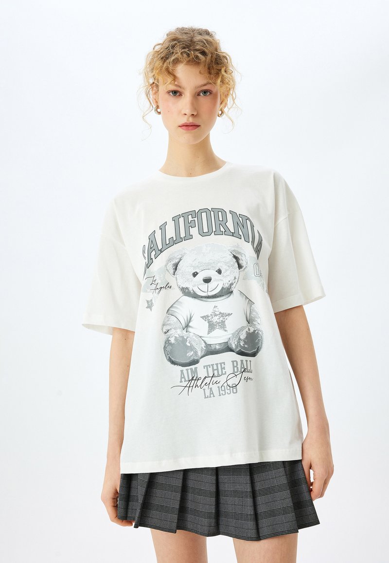 Hvid oversized t-shirt med et gråt bjørne grafik med stjerner, teksten "CALIFORNIA" og trykket "AIM THE BALL LA 1990". Glat bomuldsstof.