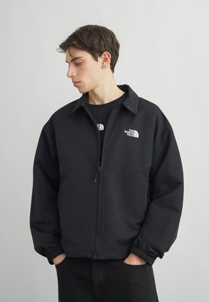 Ung mand med kort brunt hår iført en sort North Face-jakke og T-shirt, der kigger ned mod sin venstre side med hænderne i lommerne.