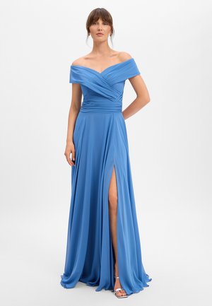 Frau trägt ein blaues, schulterfreies, langes Kleid mit hohem Schlitz und steht mit einer Hand hinter dem Rücken vor einem schlichten Hintergrund.