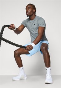 Athlete wearing a grey Nike short-sleeved shirt and light blue shorts, using a black battle rope, with white Nike shoes and wristbands.