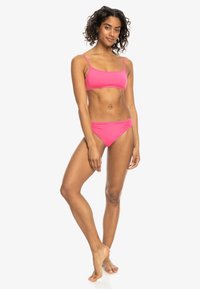 Roza bikini komplet, ki vsebuje top v stilu majice s tankimi naramnicami in srednje visokim dnom z nabornimi detajli na straneh. Gladek, elastičen material.