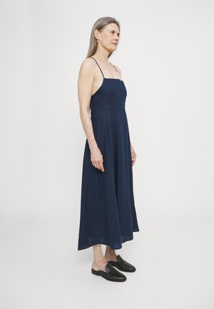 Marineblauwe mouwloze linnen jurk met een getailleerde bodice en een uitlopende, vloerlange rok; gedragen met zwarte instapschoenen; gladde textuur.