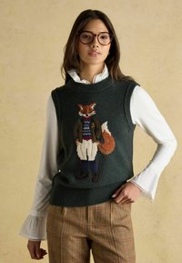 Tumši zaļš adīts bez piedurknēm svārsts ar izšūtu lapsu formālajā apģērbā. Uzvilkts virs baltas blūzes ar viļņotām piekļautēm.