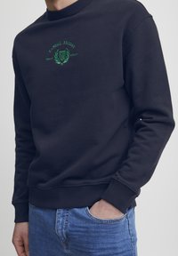 Marineblå hettegenser med ribbestrikket rund hals, med grønn brodert "CASUAL FRIDAY" logo og teksturert stoff. Kombinert med jeans.