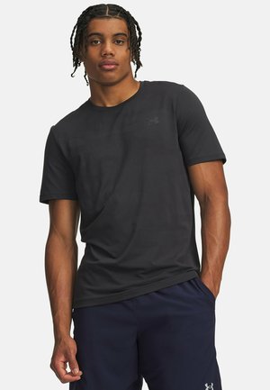 Jonge man met gevlochten haar, die een zwart Under Armour T-shirt en marineblauwe korte broek draagt, staat met één hand in de zak.