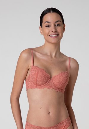 Donna sorridente con capelli scuri indossa reggiseno e slip in pizzo rosa coordinati su sfondo semplice grigio chiaro.