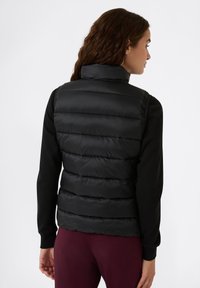 Gilet imbottito nero con sezioni a costine verticali, colletto alto e texture liscia, indossato sopra una maglia nera a maniche lunghe e leggings scuri.