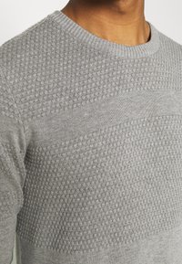 Šedý svetr s texturovaným vzorem přes hrudník a hladkým úpletem na spodní části. Má kulatý výstřih a žebrované manžety.