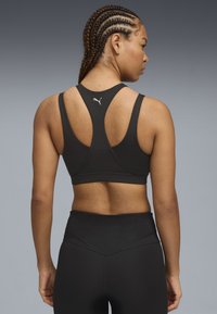 Sostenedor deportivo negro con un diseño de espalda nadadora y detalle del logo plateado en la parte posterior. La tela parece suave y elástica. Leggings negros a juego.