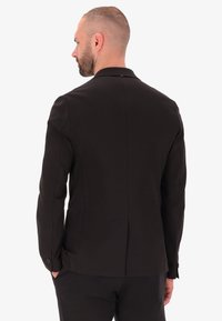 Blazer nero su misura con tessuto liscio, colletto standard e spacco posteriore singolo. Presenta polsini con bottone e due tasche laterali.