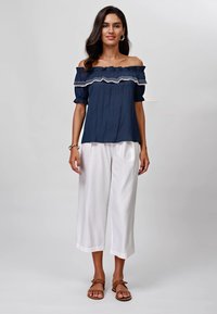 Blusa blu navy a spalle scoperte con bordi arricciati, abbinata a pantaloni bianchi a gamba larga. Il modello indossa sandali marroni. Texture lisce e tessuto morbido.
