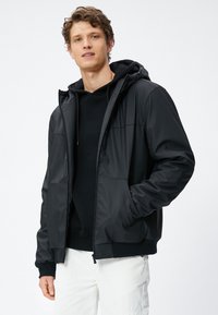 Zwarte jas met een gladde, glanzende textuur, sluiting met rits aan de voorkant en een capuchon. Heeft ribgebreide manchetten en een aansluitende taille. Gekozen over een zwarte hoodie.