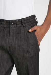Mörkgrå, texturerade byxor med dragsko i midjan, en enkel knapp och sidfickor. Materialet verkar lätt och andas.