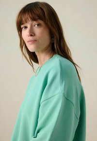Sweatshirt vert menthe avec un tissu texturé, col rond et coupe décontractée. Le modèle a les cheveux ondulés et porte des boucles d'oreilles décoratives ainsi qu'un collier fin.